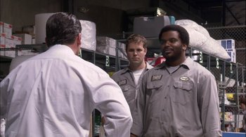 Movie still from “The Office” (2005), created by Ricky Gervais – A group of men standing next to each other in a warehouse; Medium shot, Over the shoulder angle