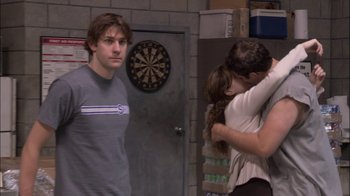 Movie still from “The Office” (2005), created by Ricky Gervais – A man and a woman hugging in a room with a man looking on; Medium shot, Over the shoulder angle