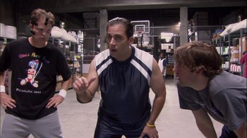 Movie still from “The Office” (2005), created by Ricky Gervais – A man standing in a room with other people; Medium shot, Over the shoulder angle