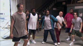Movie still from “The Office” (2005), created by Ricky Gervais – A group of men standing around in a warehouse; Wide shot, High angle