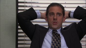 Movie still from “The Office” (2005), created by Ricky Gervais – A man in a suit and tie holding his hands behind his head; Medium shot, High angle