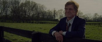 Movie still from “The Old Man & the Gun” (2018), directed by David Lowery – An older man wearing a suit and tie sitting on top of a wooden fence; Medium shot, Low angle