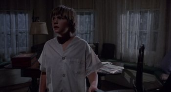 Movie still from “The Other” (1972), directed by Robert Mulligan – A boy in a white shirt is standing in a living room; Medium shot, Low angle