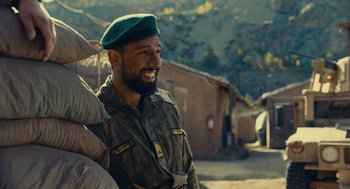 Movie still from “The Outpost” (2019), directed by Rod Lurie – A man in a green hat smiles for the camera; Medium shot, Over the shoulder angle