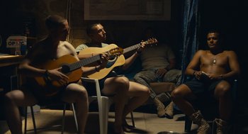 Movie still from “The Outpost” (2019), directed by Rod Lurie – A group of people sitting in a room with guitars; Wide shot, Over the shoulder angle