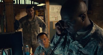 Movie still from “The Outpost” (2019), directed by Rod Lurie – A man in fatigues talking on a phone while two other men watch; Medium shot, Over the shoulder angle