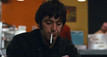 Movie still from “The Panic in Needle Park” (1971), directed by Jerry Schatzberg – A man sitting at a table with a cigarette in his mouth; Close Up shot, Over the shoulder angle