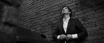 Movie still from “The Party” (2017), directed by Sally Potter – A black and white photo of a man standing in front of a brick wall; Medium shot, Low angle