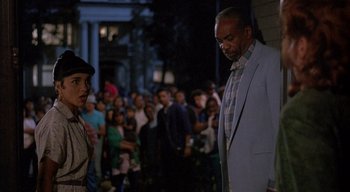 Movie still from “The People Under the Stairs” (1991), directed by Wes Craven – A crowd of people standing in front of a building at night; Medium shot, Over the shoulder angle