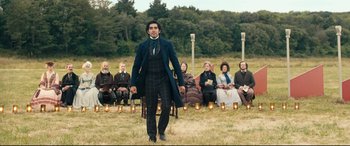 Movie still from “The Personal History of David Copperfield” (2019), directed by Armando Iannucci – A group of people sitting in a field with candles in front of a man; Wide shot, Over the shoulder angle