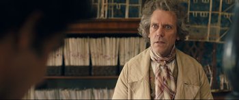 Movie still from “The Personal History of David Copperfield” (2019), directed by Armando Iannucci – An older man in a jacket and a neck tie in front of a shelf of papers; Close Up shot, Over the shoulder angle