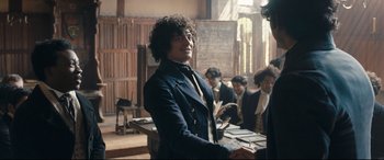 Movie still from “The Personal History of David Copperfield” (2019), directed by Armando Iannucci – A man standing in front of a group of people; Medium shot, Over the shoulder angle