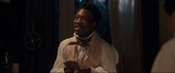 Movie still from “The Personal History of David Copperfield” (2019), directed by Armando Iannucci – A man wearing a vest and bow tie holding a cell phone; Medium shot, Over the shoulder angle