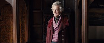 Movie still from “The Personal History of David Copperfield” (2019), directed by Armando Iannucci – An older man wearing a red coat and a purple vest; Medium shot, Over the shoulder angle