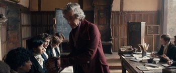 Movie still from “The Personal History of David Copperfield” (2019), directed by Armando Iannucci – An older man wearing a red coat is standing in front of a chalkboard; Medium shot, Over the shoulder angle