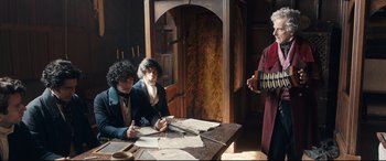 Movie still from “The Personal History of David Copperfield” (2019), directed by Armando Iannucci – Two people sitting at a table with papers and a man playing an accordion; Medium shot, High angle