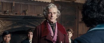 Movie still from “The Personal History of David Copperfield” (2019), directed by Armando Iannucci – An older man in a red coat standing in front of a group of young men; Medium shot, Over the shoulder angle
