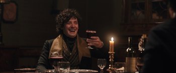 Movie still from “The Personal History of David Copperfield” (2019), directed by Armando Iannucci – A man sitting at a dinner table holding a glass of wine; Medium shot, Over the shoulder angle