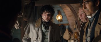 Movie still from “The Personal History of David Copperfield” (2019), directed by Armando Iannucci – A young man in a coat and tie standing next to a light fixture; Medium shot, Over the shoulder angle