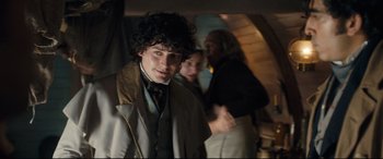 Movie still from “The Personal History of David Copperfield” (2019), directed by Armando Iannucci – A young man in a coat and tie standing in front of two other people; Medium shot, Over the shoulder angle