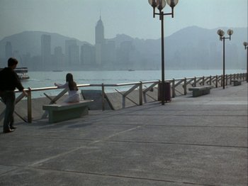 Movie still from “The Pillow Book” (1995), directed by Peter Greenaway – A woman sitting on a bench near a body of water; Extreme Wide shot, High angle