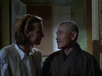 Movie still from “The Pillow Book” (1995), directed by Peter Greenaway – A man standing next to another man in front of a wall; Close Up shot, Over the shoulder angle