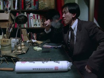 Movie still from “The Pillow Book” (1995), directed by Peter Greenaway – A man sitting at a table smoking a cigarette in front of another man; Medium shot, High angle