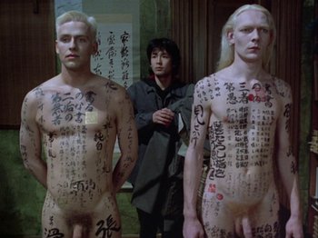 Movie still from “The Pillow Book” (1995), directed by Peter Greenaway – A man standing in front of two naked men with chinese writing on them; Medium shot, High angle