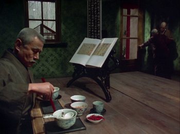Movie still from “The Pillow Book” (1995), directed by Peter Greenaway – An old man sitting in front of an open book on the floor; Wide shot, High angle