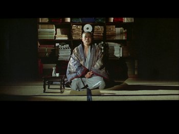 Movie still from “The Pillow Book” (1995), directed by Peter Greenaway – A woman sitting on the floor in front of a stack of books; Wide shot, Low angle