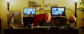 Movie still from “The Prince” (2014), directed by Brian A. Miller – A man sitting at a desk in front of two computer monitors; Medium shot, Over the shoulder angle