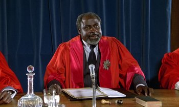 Movie still from “The Professional” (1981), directed by Georges Lautner – A man sitting at a table wearing a red robe; Medium shot, Low angle