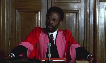 Movie still from “The Professional” (1981), directed by Georges Lautner – A man in a red and pink robe sitting in front of a microphone; Close Up shot, Low angle