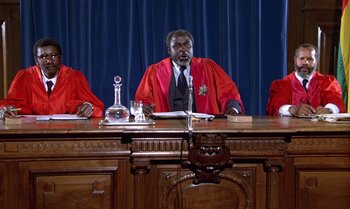 Movie still from “The Professional” (1981), directed by Georges Lautner – A judge sitting at a table in front of two other judges; Medium shot, Low angle
