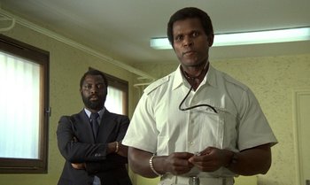 Movie still from “The Professional” (1981), directed by Georges Lautner – Two men standing next to each other in a room; Medium shot, Low angle