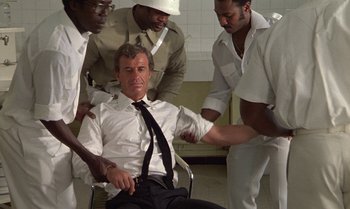 Movie still from “The Professional” (1981), directed by Georges Lautner – A group of men standing next to a man in a chair; Medium shot, High angle