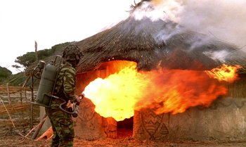 Movie still from “The Professional” (1981), directed by Georges Lautner – A man in camouflage is standing in front of a burning hut; Wide shot, Low angle
