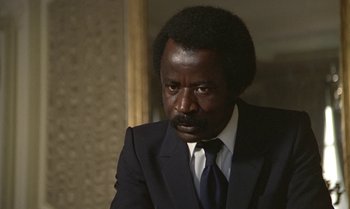 Movie still from “The Professional” (1981), directed by Georges Lautner – A man wearing a suit and tie sitting in a room; Close Up shot, High angle