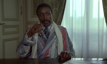 Movie still from “The Professional” (1981), directed by Georges Lautner – A man sitting at a table with a tie around his neck; Medium shot, Low angle