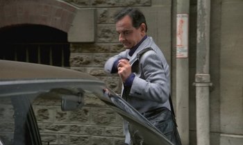 Movie still from “The Professional” (1981), directed by Georges Lautner – A man in a gray jacket and a blue tie is standing next to a car; Medium shot, High angle