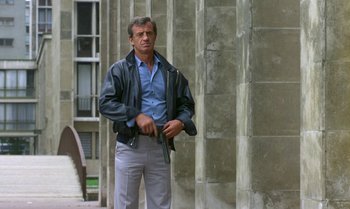 Movie still from “The Professional” (1981), directed by Georges Lautner – A man standing in front of a building holding a gun; Medium shot, Low angle