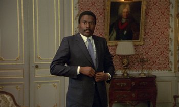 Movie still from “The Professional” (1981), directed by Georges Lautner – A man in a suit and tie standing in front of a painting; Medium shot, Low angle