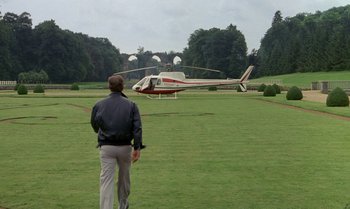Movie still from “The Professional” (1981), directed by Georges Lautner – A man standing in front of a helicopter on a field; Wide shot, Over the shoulder angle