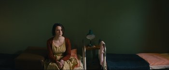 Movie still from “Journal 64” (2018), directed by Christoffer Boe – A woman sitting on a bed next to a lamp; Medium shot, High angle