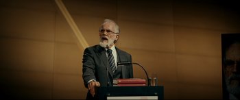 Movie still from “Journal 64” (2018), directed by Christoffer Boe – An older man in a suit is speaking at a podium; Medium shot, Over the shoulder angle