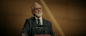 Movie still from “Journal 64” (2018), directed by Christoffer Boe – An older man in a suit is speaking at a podium; Medium shot, Over the shoulder angle