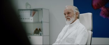 Movie still from “Journal 64” (2018), directed by Christoffer Boe – An older man in a white lab coat sitting in front of a glass case; Medium shot, Over the shoulder angle