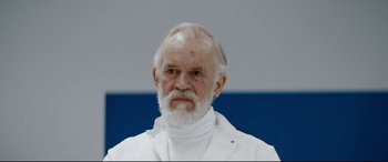 Movie still from “Journal 64” (2018), directed by Christoffer Boe – An older man with a white beard wearing a lab coat; Close Up shot, Low angle