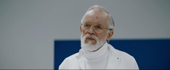 Movie still from “Journal 64” (2018), directed by Christoffer Boe – An older man with a beard and glasses wearing a white lab coat; Close Up shot, Over the shoulder angle