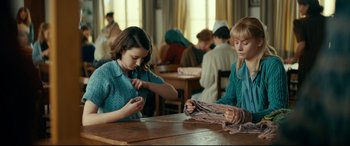 Movie still from “Journal 64” (2018), directed by Christoffer Boe – Two young girls sitting at a wooden table knitting; Medium shot, High angle
